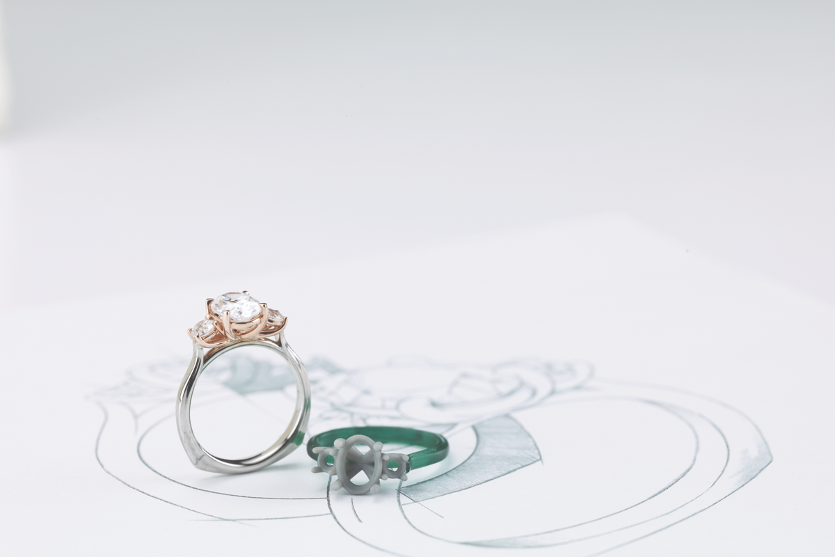 A custom-made ring with the CAD prototype laying beside it and the original hand-drawn design in the background.