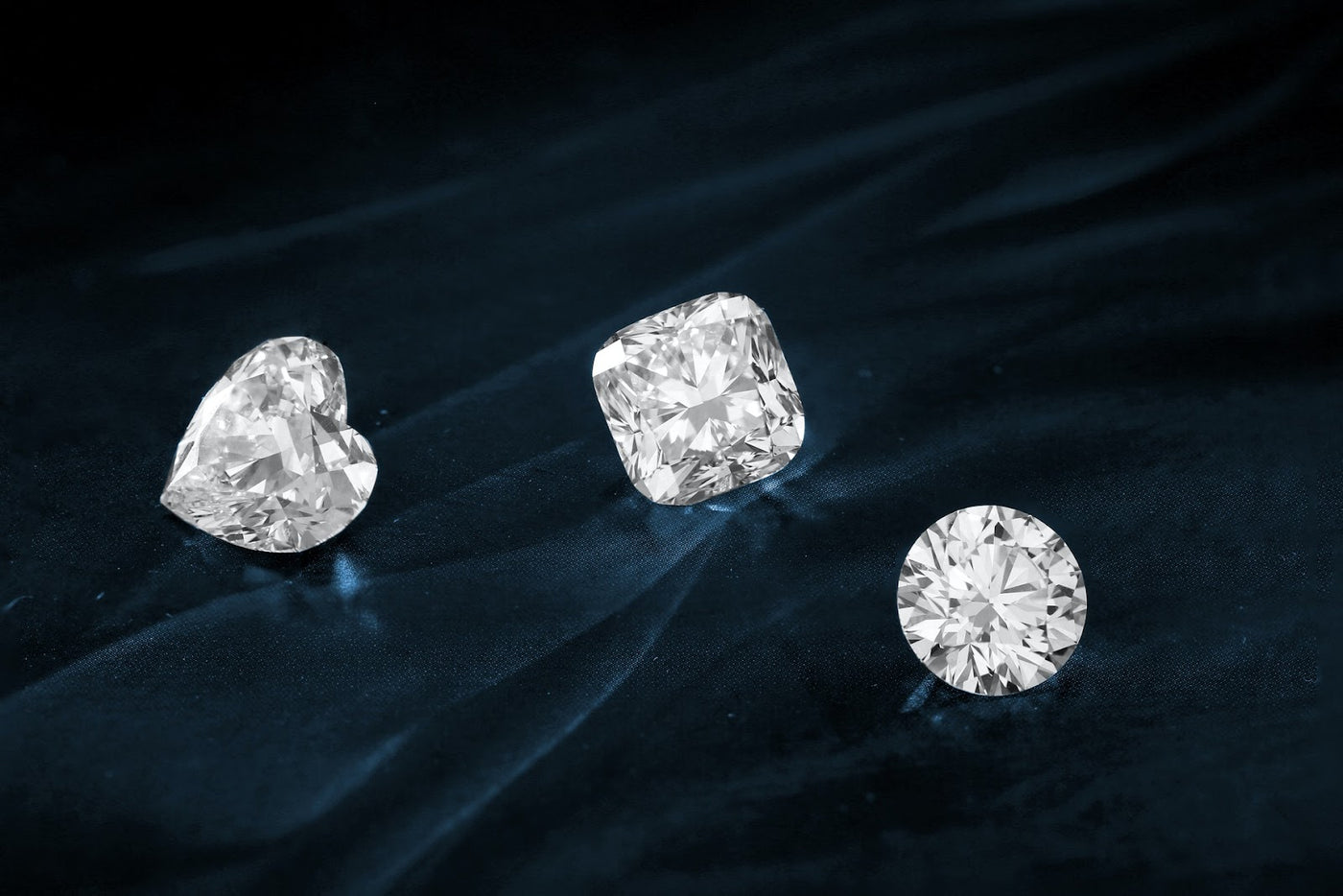 Three differently cut lab-grown diamonds, one heart shaped, one round and one cushion cut on a black background for the 4 C's of diamond grading.