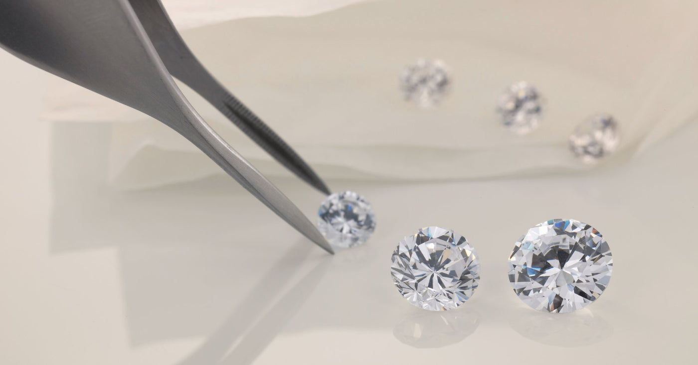 Four loose lab grown luxury diamonds of different cuts and shapes laid out on a black surface with one being picked up by jewelry tweezers.