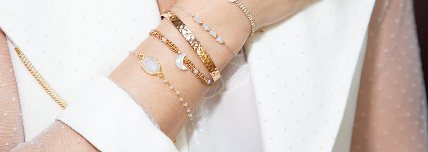 Close up photo of a women's wrist with a stack of different gold bangles, and chain bracelets featuring lab-grown diamonds, with her classy white dress in the background