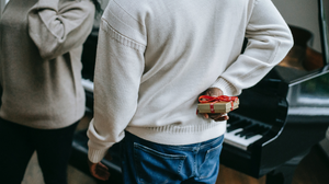 Two people standing in front of a piano during the holidays with one person holding a gold box, with red ribbon around it as a gift.