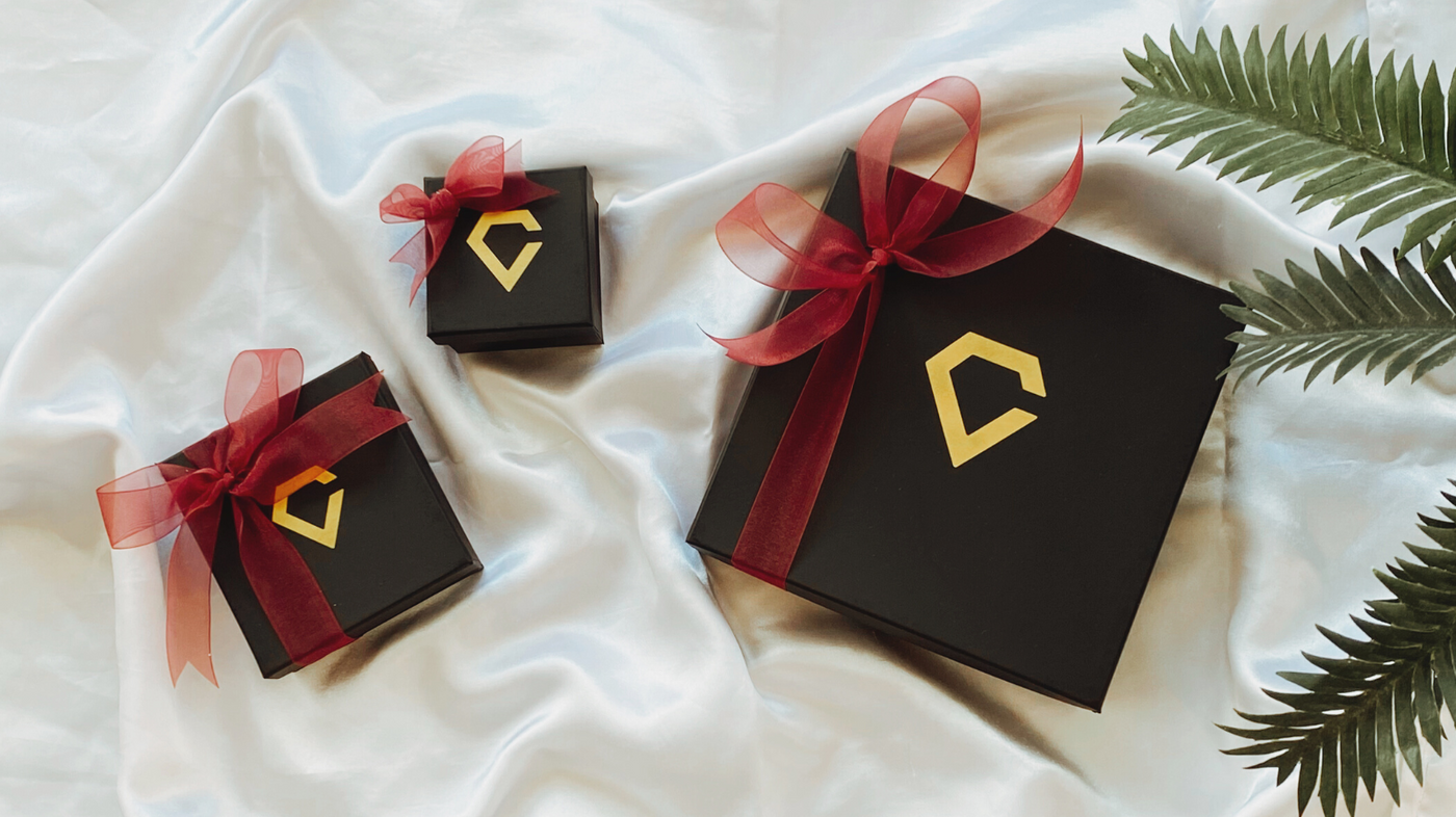 Three black boxes with red ribbon around them and the gold Carbon Diamonds logo on them, all boxes sitting on a white cloth with greenery on the side.