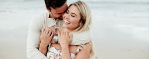 A couple recently engaged, he hugs her from behind on a beach with her sustainable diamond engagement ring shown off on her hand.